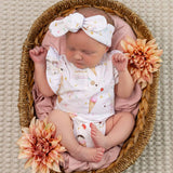 A sleeping newborn baby wearing a white short-sleeve bodysuit with a pastel ice cream cone print and a matching bow headband, posed in a woven basket with pink fabric lining and peach flowers.