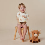 A baby sitting on a small wooden stool wearing a cream bib with a brown cow pattern, posed next to a plush brown cow toy against a solid beige background.