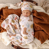 A sleeping newborn baby swaddled in a western-print blanket and wearing a matching hat, resting in a wicker basket with brown and cream knit blankets.