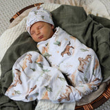 A sleeping newborn baby swaddled in a white blanket with a brown fox and cactus print, wearing a matching white hat, resting in a wicker basket lined with a dark green blanket.