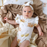A baby lying on cream and brown textured blankets, wearing a white short-sleeve bodysuit with a yellow and brown animal print and a matching white headband.