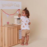 A toddler stands barefoot in front of a wooden play cafe setup, wearing a white short-sleeve t-shirt with a light brown teddy bear print and beige shorts. The scene includes a sign reading 'Snuggle Café' and a small toy coffee machine.