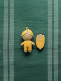 A small yellow doll-shaped accessory and a matching yellow shield-shaped accessory displayed on a green striped fabric background.
