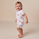 A smiling toddler wearing a white short-sleeve romper with a brown animal print and beige trim, standing barefoot against a plain beige studio background.