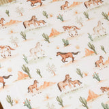 A close-up of fabric featuring a repeating western desert print with brown and white horses, cacti, sandy hills, and small sun motifs on an off-white background.