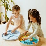 Two young girls playing with a sensory bin filled with water and small toys on a light-colored surface.