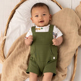 A baby lying on a beige muslin blanket inside a wicker basket, wearing an olive green overall romper over a white short-sleeve shirt with colorful cartoon prints.