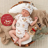 A sleeping newborn baby wearing a white Christmas-themed short-sleeve bodysuit and matching hat, lying in a wicker basket with a beige knit blanket, a red round sticker that says 'MY FIRST Christmas,' and a knitted reindeer toy.