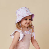 A young child wearing a light pink sun hat with a purple floral pattern and a matching pink overall dress, set against a solid beige background.
