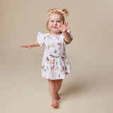 A toddler girl wearing a white ruffle-sleeve dress with a western print of horses, cacti, and a brown belt, walking barefoot against a solid beige background.