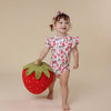Child wearing a strawberry-themed outfit holding a large plush strawberry against a beige background