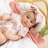 A baby lying on a white blanket in a wicker basket, wearing a light pink short-sleeve romper with a bunny print and a matching pink bow headband.
