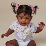 A baby wearing a white ribbed bodysuit and a white floral bib with pink, purple, and green botanical prints. The baby has dark hair styled in two pigtails with pink bows, sitting against a solid beige background.