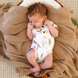 A sleeping newborn baby wearing a white short-sleeve bodysuit with a desert animal and cactus print, lying on a textured tan blanket inside a wicker basket.