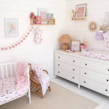 A styled nursery room with white shiplap walls, a white dresser with a floral changing pad, a white crib with matching bedding, a rattan rocker with a pink blanket, and pink decorative accents including a pom-pom garland and framed wall art.