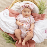 A baby doll or realistic baby toy wearing a light pink short-sleeve romper with small animal prints and a matching bow headband, posed in a woven basket with white and pink blankets and greenery.
