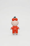 A small stylized figurine of a child wearing a bright red jumpsuit and a red bow with a green stem, standing against a plain light gray background.