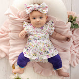 A baby lying on a pink ruffled blanket wearing a white floral dress with ruffled sleeves, a matching floral bow headband, and purple leggings.