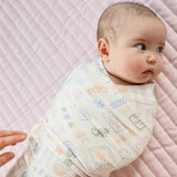 A baby swaddled in a soft, patterned muslin swaddle blanket, lying on a light pink quilted blanket. The swaddle has a cream base with a pastel multicolor print of animals, flowers, and geometric shapes.
