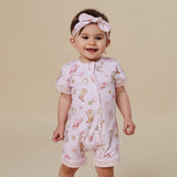 A smiling baby girl wearing a light pink short-sleeve zip-up romper with a cowboy boot and hat pattern, paired with a matching pink bow headband, set against a solid beige background.