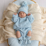 Newborn baby in a blue knitted outfit with pom-pom details, lying on a textured blanket.