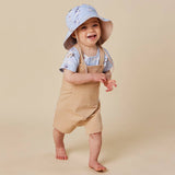 A smiling toddler standing barefoot on a warm beige studio background, wearing a light blue patterned sun hat, a light blue short-sleeve shirt, and tan overall-style overalls.