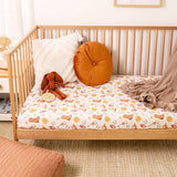 A wooden crib or toddler bed with a fitted sheet featuring a playful multicolor horse and bit pattern, styled with an orange round pillow, a white textured pillow, a plush bunny, and a brown throw blanket.