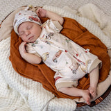 A sleeping newborn baby wearing a cream-colored zip-up sleeper and matching hat with a vintage car pattern, nestled in a rust-colored muslin wrap on a white knit blanket.