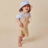 A smiling toddler wearing a light blue patterned sun hat, a light blue short-sleeve shirt, and beige overall-style baby shorts, posed against a plain warm beige studio background.