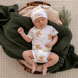 A sleeping newborn baby wearing a white short-sleeve romper and matching hat with teddy bear prints, lying in a wicker basket on a beige textured blanket with a dark green knit throw.
