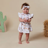 A toddler stands barefoot in a studio setting wearing a white ruffled dress with a western horse print and a matching white bow headband. The background is a solid beige with decorative props, including a cactus on the left and a bale of hay on the right.
