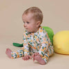 Baby wearing a lemon-patterned onesie sitting on a beige surface with green and yellow pillows.