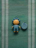 A small blue and yellow character keychain or accessory next to a round blue case with a matching loop, placed on a green striped fabric background.
