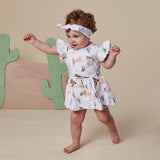 A toddler girl wearing a white ruffle-sleeve dress and matching headband with a desert animal print, standing barefoot in front of a beige backdrop with a wooden cactus decoration.