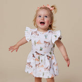 A toddler girl with blonde hair in a ponytail and a pink bow, wearing a white short-sleeve dress with a western desert print of horses, cacti, and mountains. She is standing against a solid beige background with her arms outstretched.