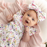 A baby lying on a soft pink blanket, wearing a white floral romper with ruffled sleeves and a matching bow headband.