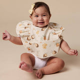 A baby sitting on a beige studio floor wearing a cream bib with a bee and honeycomb print, along with a matching cream bow in their hair.