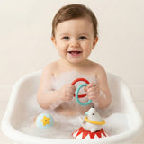 A smiling baby sitting in a white bathtub filled with soapy water, holding colorful toy rings. A white polar bear bath toy is visible in the tub.