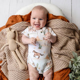 A smiling baby lying on their back, wearing a white short-sleeve bodysuit with a desert safari print of cacti and horses. The baby is surrounded by cozy props, including a textured beige knit blanket, an orange throw blanket, a plush toy, and a white wooden background.