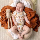 A smiling baby lies on a round beige lounger, wearing a cream short-sleeve bodysuit with a colorful food-themed print. The baby is surrounded by a chunky rust-orange knit blanket and a beige plush toy, set against a textured cream background.