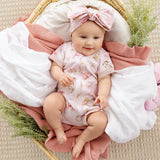 A baby wearing a light pink short-sleeve romper with a bunny print and a matching bow headband, lying on a pink ribbed blanket inside a woven basket with white and pink blankets.