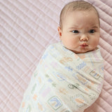 A baby swaddled in a light cream muslin swaddle blanket with a colorful pastel pattern of sea creatures, shown lying on a pink quilted mattress.