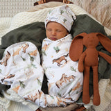 A sleeping newborn baby swaddled in a white desert-themed blanket with cacti and horses, wearing a matching hat, nestled in a basket with a brown plush bunny toy.