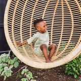 Child sitting in a large wicker chair outdoors with plants around
