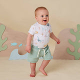 A baby standing barefoot in a studio setting, wearing a white short-sleeve top with a colorful ocean-themed print and mint green shorts with a white drawstring. The background features a beige backdrop with decorative coral and wave cutouts.