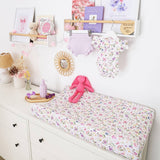 A baby changing table covered with a white floral changing pad, styled in a nursery with a pink stuffed bunny, floral baby clothes hanging on a wooden rack, and framed decor on a white wall.