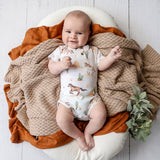 A baby lying on a white cushion, wrapped in a chunky beige knit blanket and rust-colored fabric, wearing a white short-sleeve bodysuit with a desert animal print. A small green plant is placed to the right on a white wood background.