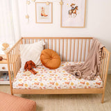 A light natural wood crib with a white mattress covered in a cowboy boot and hat print, styled with a white pillow, an orange round cushion, a brown stuffed animal, and a beige ruffled throw blanket. The crib is set in a nursery with a woven rug, a side table, and framed western-themed art on the wall.