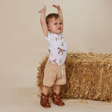 A toddler stands with arms raised in front of a hay bale, wearing a white short-sleeve shirt with a western horse print, beige shorts, and brown cowboy boots against a beige studio backdrop.