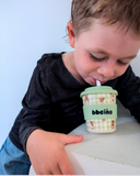 Child drinking from a green sippy cup with a checkered pattern and bear design, featuring the brand 'bbeino'.